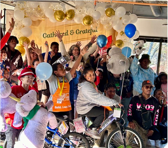 Children and volunteers celebrating with new bicycles, stuffed animals, and balloons beneath a balloon arch