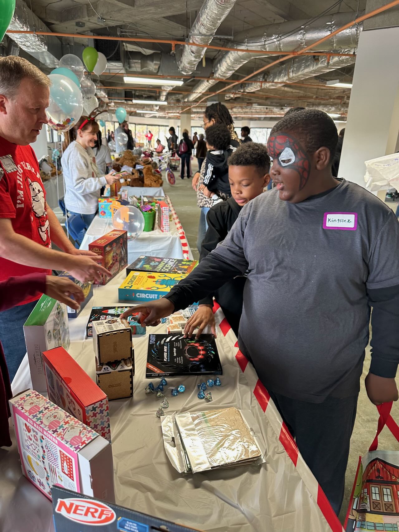 Children at a table of boxed toys and craft kits while a volunteer helps