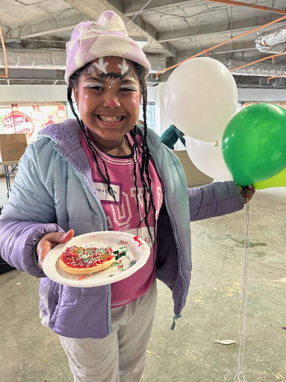 A smiling girl with reindeer face paint holds a decorated cookie and a bunch of balloons.