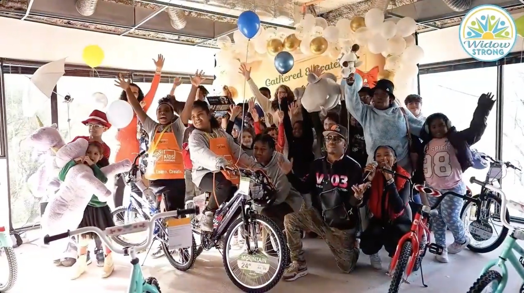 Children and volunteers celebrating together with new bicycles and stuffed animals beneath a balloon arch at the Merry Widows Gift Gathering