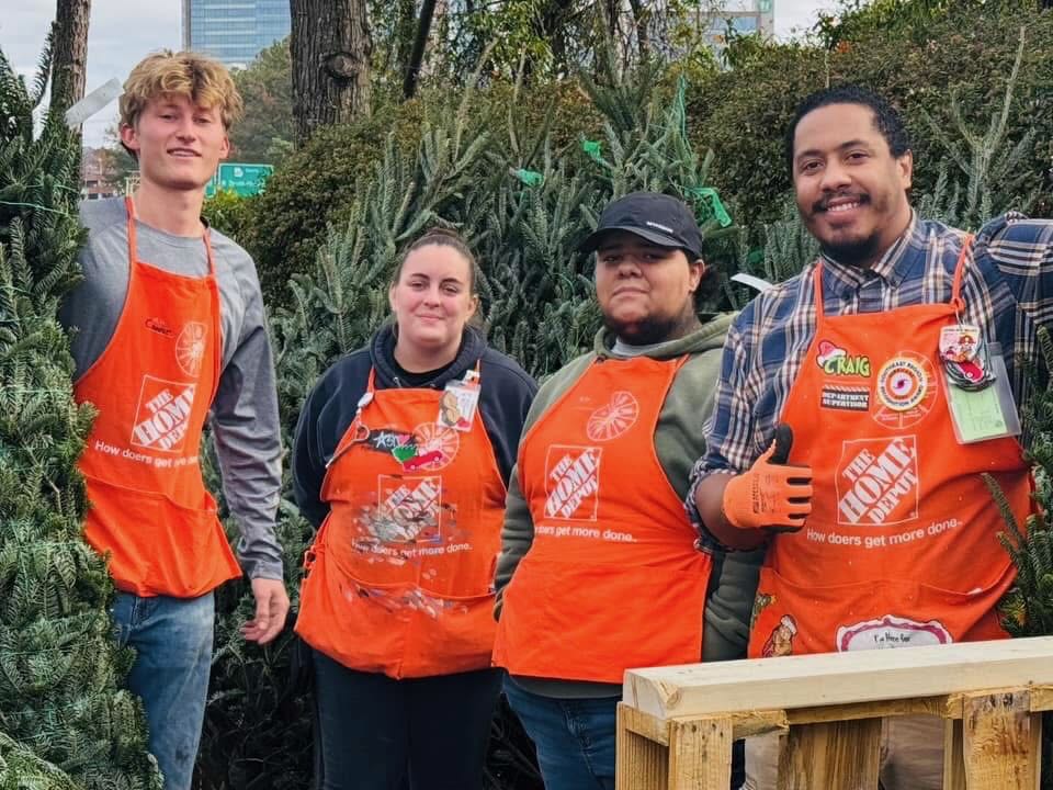 Four Home Depot associates in orange aprons posing among donated Christmas trees