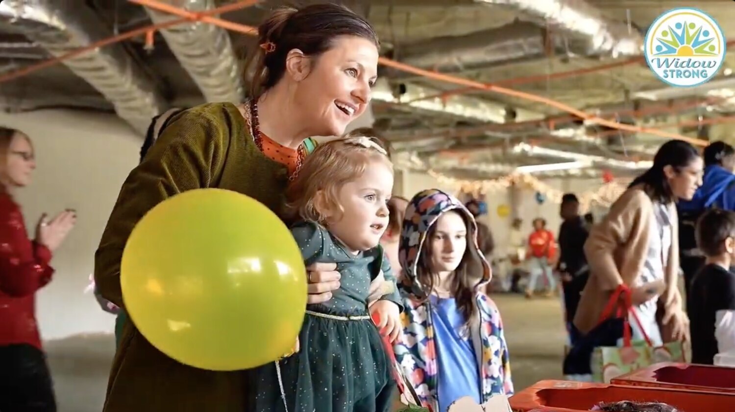 A smiling woman holding a toddler in a sparkly green dress beside a large lime-green balloon, with an older girl in a floral jacket standing close by